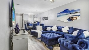 A modern living room with a large blue sectional sofa, a ceiling fan, and a beach-themed wall art. - Luxury n cozy 3 bed 2 bath vacation property in a gated community (Kissimmee)