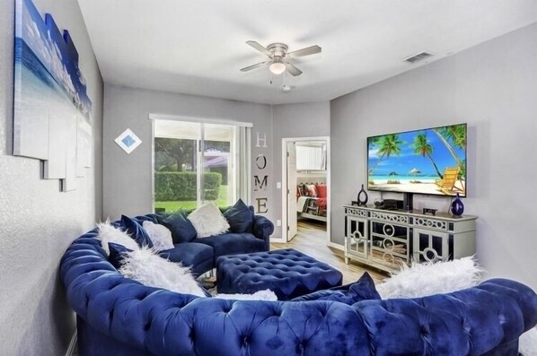 Una sala de estar con un sofá azul curvo, un ventilador de techo, un televisor y un cuadro de una escena de playa.