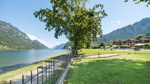 A lakeside view with a large tree, a grassy area, and a building with a red roof. - Apartment by Lake Idro with Mountain Views (Idro (bs))