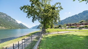 A lakeside property with a grassy lawn, a wooden fence, and a building with a red roof. - Apartment on Lake Idro with Pool (Idro (bs))