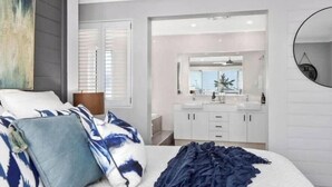 A bedroom with a bed, pillows, a bedside table, a mirror, and a bathroom with a sink and cabinets.