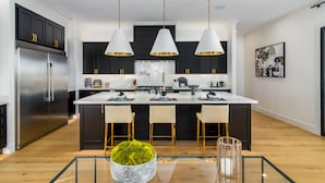 A modern kitchen with dark cabinets, a stainless steel refrigerator, a central island with bar stools, pendant lighting, and a glass-top dining table.