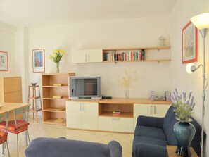 Una sala de estar con televisor, estanterías, un sofá, una mesa de comedor con sillas y un jarrón con flores.