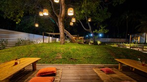 Un patio al aire libre con bancos de madera y cojines, rodeado de árboles e iluminado con luces colgantes.