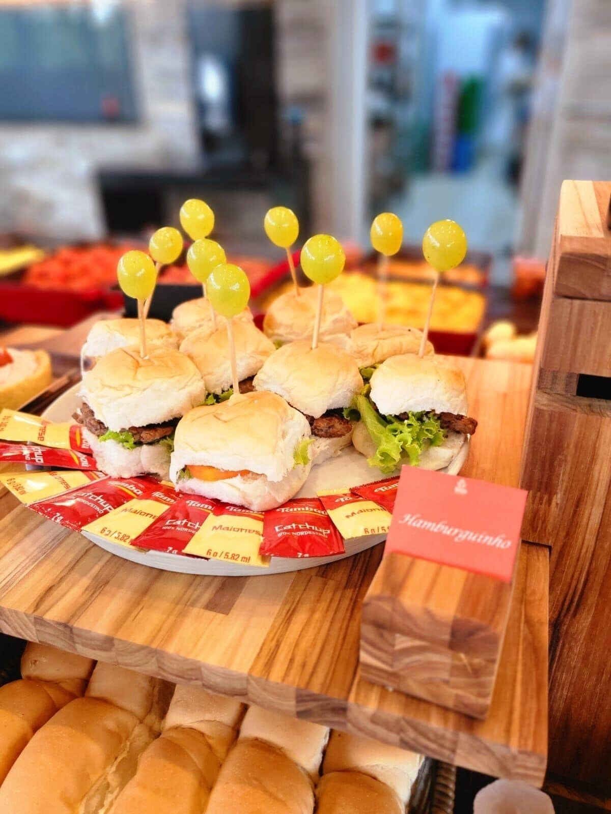 A display of mini sandwiches with yellow toothpicks, surrounded by packaged condiments and a sign indicating 'Hamburgutinho'.