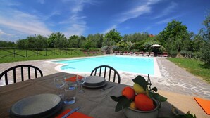 A dining table set with plates, glasses, and a bowl of fruit, overlooking a swimming pool with a garden and outdoor seating area.