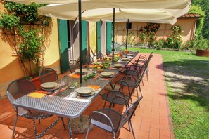 Un lungo tavolo da pranzo all'aperto con sedie in metallo, immerso in un giardino e affiancato da un edificio con persiane verdi.