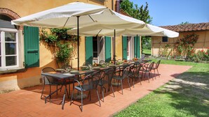 An outdoor dining area with a long table and chairs, covered by large umbrellas, set against a building with green shutters and a tiled roof.