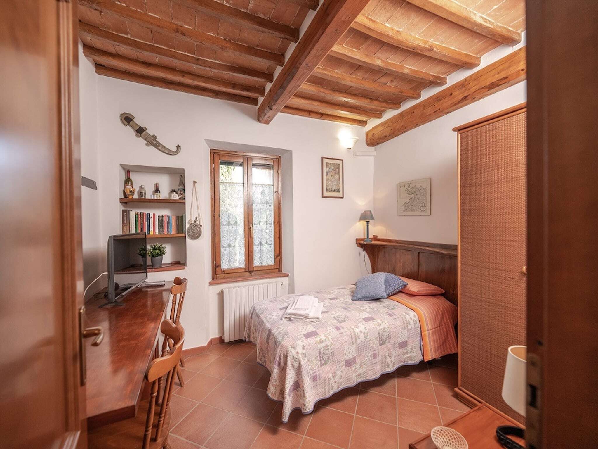 A cozy bedroom with a wooden ceiling, a bed with a patterned bedspread, a bookshelf, a radiator, and a window with lace curtains.