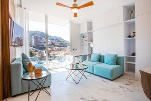 En moderne stue med sofa, sofabord og tv. Der er en balkon med udsigt til bygninger og bakker.