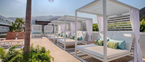 A rooftop lounge area with white canopies, cushioned seating, and a view of the mountains.