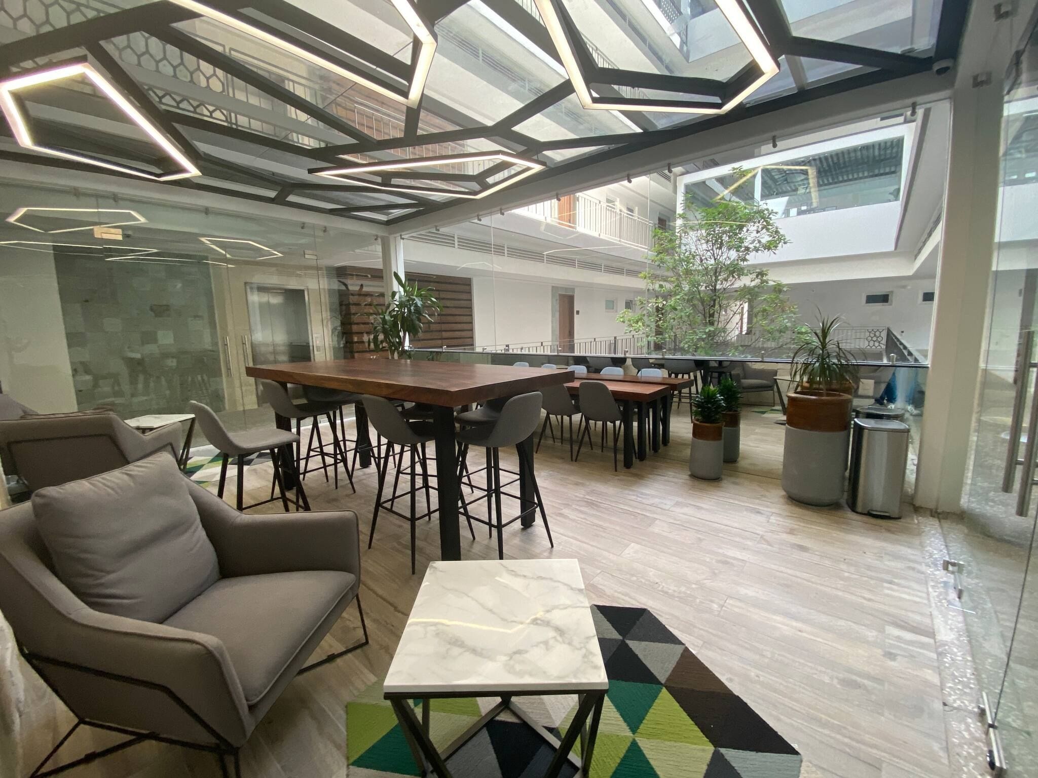 Ein modernes Interieur mit hoher Decke, einem zentralen Holztisch und umliegenden Stühlen sowie einer Glaskabine.
