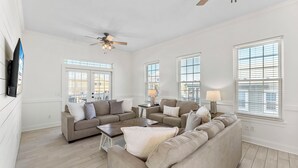 Ein geräumiges Wohnzimmer mit einem beigefarbenen Ecksofa, einem Couchtisch und einem Deckenventilator. Große Fenster mit Jalousien und ein an der Wand befestigter Flachbildfernseher.