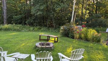 Un jardín con un hogar para fuego, un banco de madera y sillas tipo Adirondack.