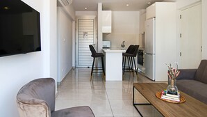 A modern living space with a flat-screen TV mounted on the wall, a wooden coffee table with books and a vase, a grey sofa, and a kitchen area with a fridge and a small island with bar stools. - Phaedrus Living: Seaside Executive Flat Harbour 207 (Paphos)