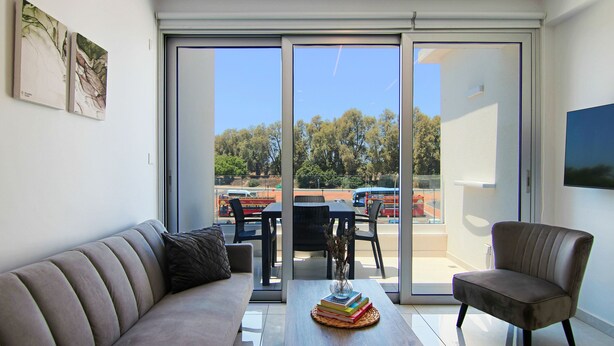 A modern living room with a sofa, a coffee table with books, a chair, and a view through a sliding glass door to an outdoor area with a table and chairs. - Phaedrus Living: Seaside Executive Flat Harbour 207 (Paphos)