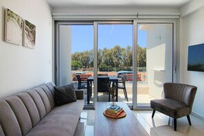 A modern living room with a sofa, a coffee table with books, a chair, and a view through a sliding glass door to an outdoor area with a table and chairs. - Phaedrus Living: Seaside Executive Flat Harbour 207 (Paphos)