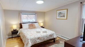 A bedroom with a bed, two bedside lamps, a window with curtains, a framed picture on the wall, and a wooden desk with a remote control. - Winter Lore (Rutland)