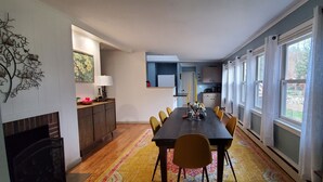 A dining area with a long table, chairs, a fireplace, a kitchen in the background, and large windows. - Winter Lore (Rutland)
