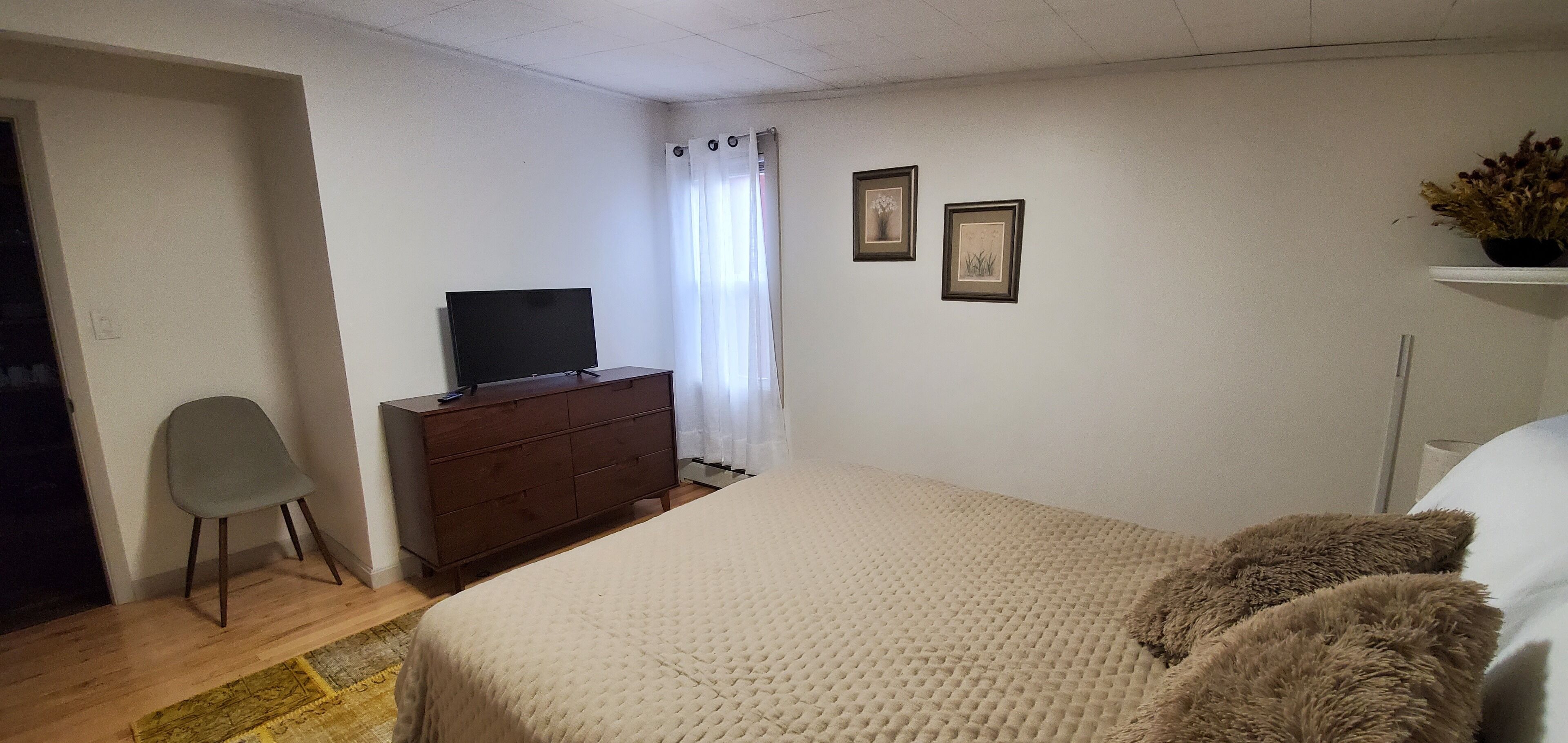 Ein Schlafzimmer mit einem Bett, einem Schminktisch, einem Sessel, einem Fernseher und zwei gerahmten Bildern an der Wand.