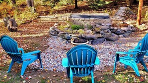 A stone fire pit surrounded by rocks and foliage, with three blue chairs arranged around it. - Winter Lore (Rutland)
