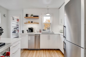A modern kitchen with white cabinetry, a stainless steel dishwasher, and a built-in oven. There are shelves above the sink holding various items, and a window above the sink provides natural light. - MINNeSTAY* Nokomis Blue | Quiet (Minneapolis)