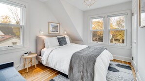 Een slaapkamer met een bed, twee bedlampen, een ingelijst foto aan de muur en een raam met uitzicht op bomen.