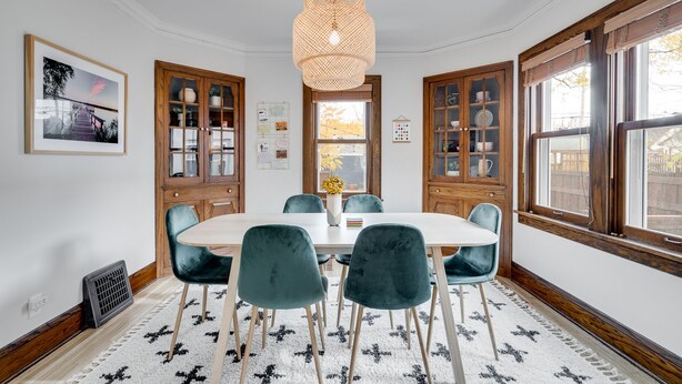 Een eetkamer met een ronde tafel, zes stoelen, een hanglamp, houten kasten en een raam met uitzicht.