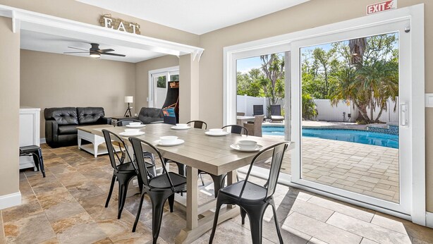 Un comedor con una mesa dispuesta para cuatro, un sofá de cuero negro y una piscina visible a través de la puerta de cristal.