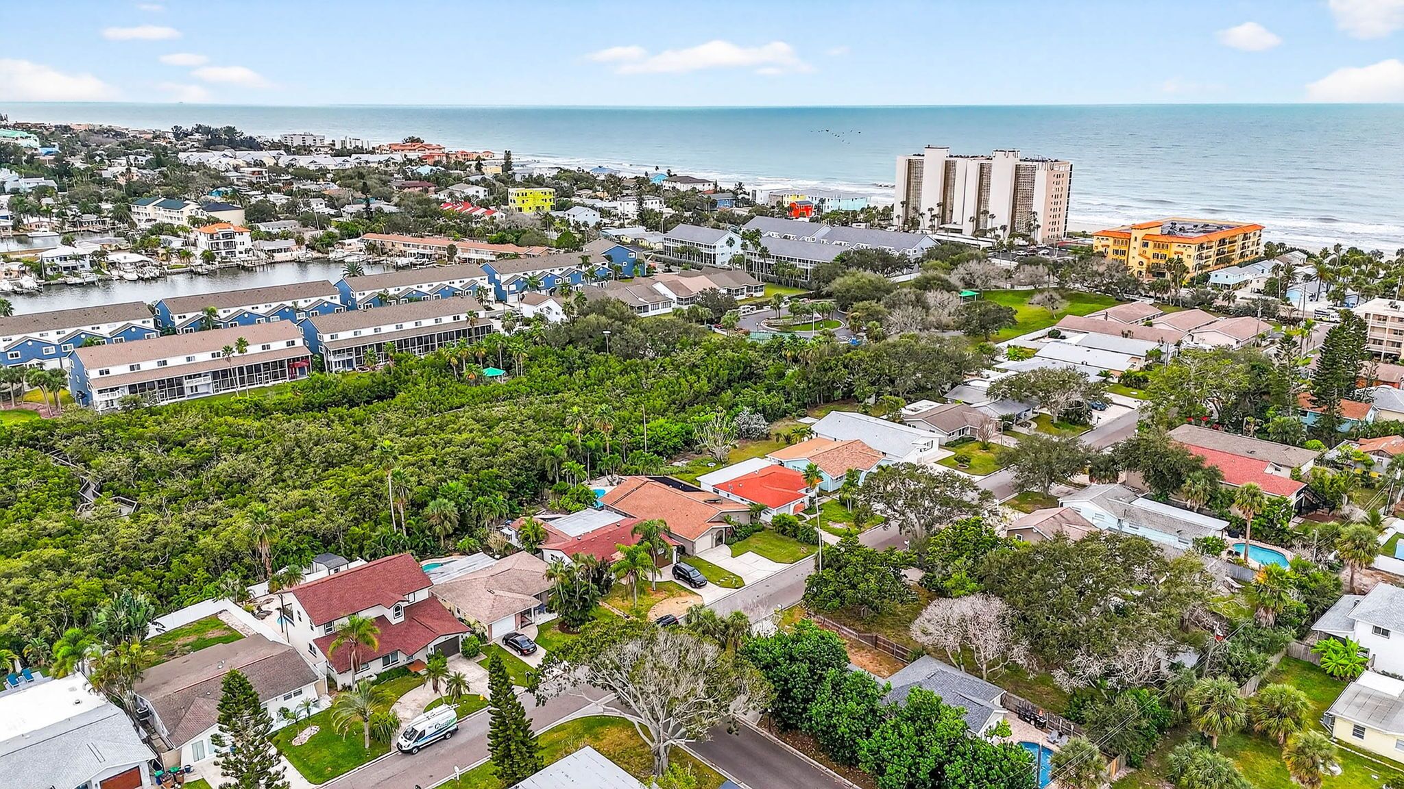 An aerial view of a coastal residential area with houses, a marina, and a hotel with multiple stories and balconies overlooking the beach.