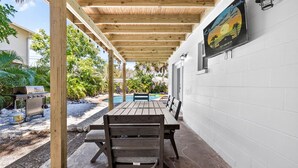 An outdoor patio with wooden tables and chairs, a barbecue grill, and a pool in the background. - Heated Pool/2 min walk 2 the Beach/Sleeps 9/Cute (Indian Rocks Beach)