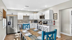 A modern kitchen with white cabinets, a stainless steel refrigerator, a microwave, and a wooden dining table with blue chairs. - Heated Pool/2 min walk 2 the Beach/Sleeps 9/Cute (Indian Rocks Beach)