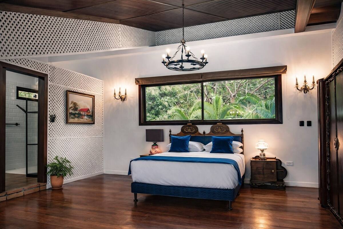 Een slaapkamer met een groot raam, een bed met blauw beddengoed, een kroonluchter en een plant in een pot.