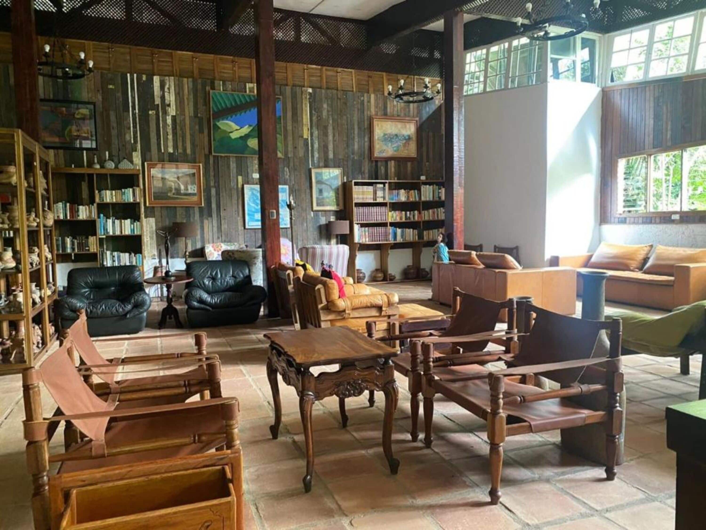 Una habitación amplia con una gran mesa de madera, varias opciones de asientos que incluyen un sofá y sillas, una estantería llena de libros y un ventanal que proporciona luz natural.