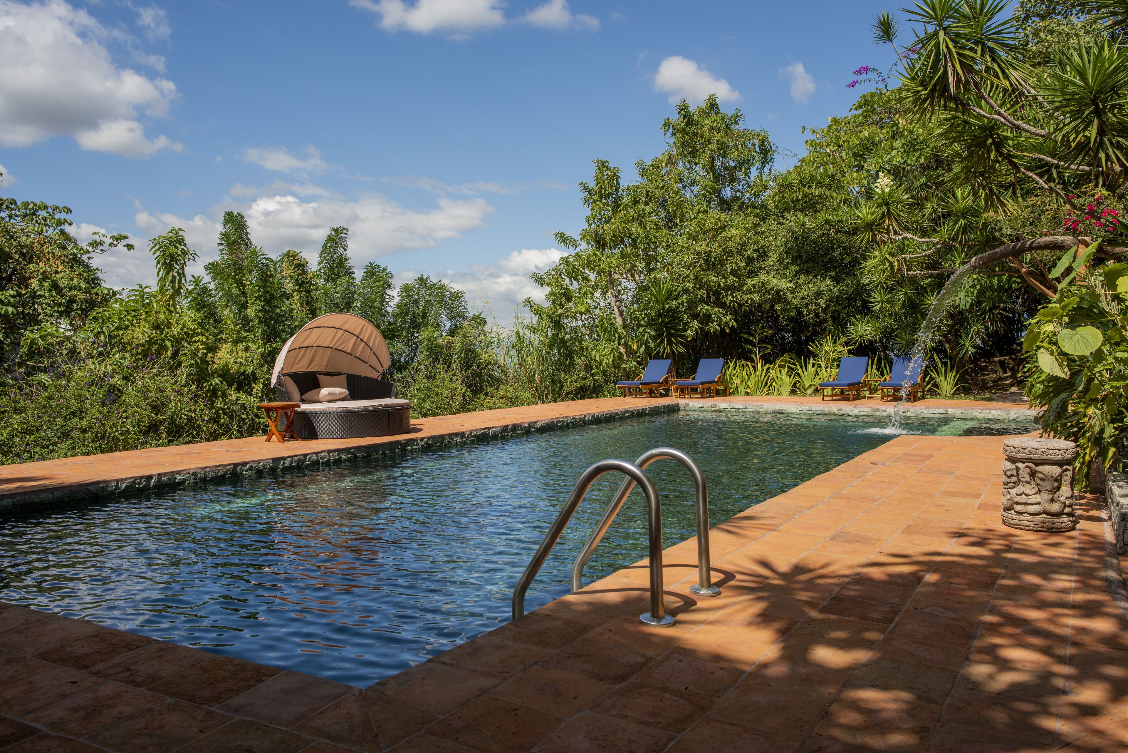A swimming pool with a sunken seating area, surrounded by lush greenery and a clear sky.