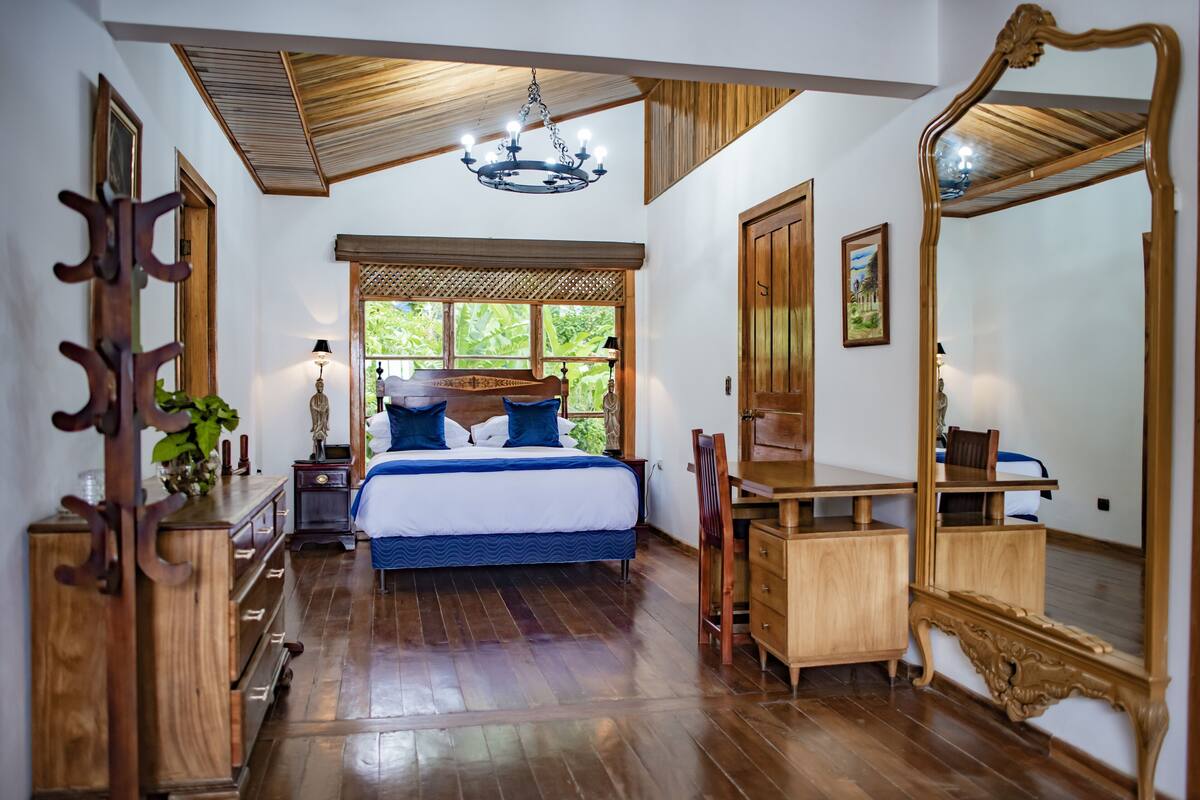 Een slaapkamer met een houten vloer, een bed met blauw beddengoed, een bureau, een stoel, een grote spiegel en een kroonluchter.