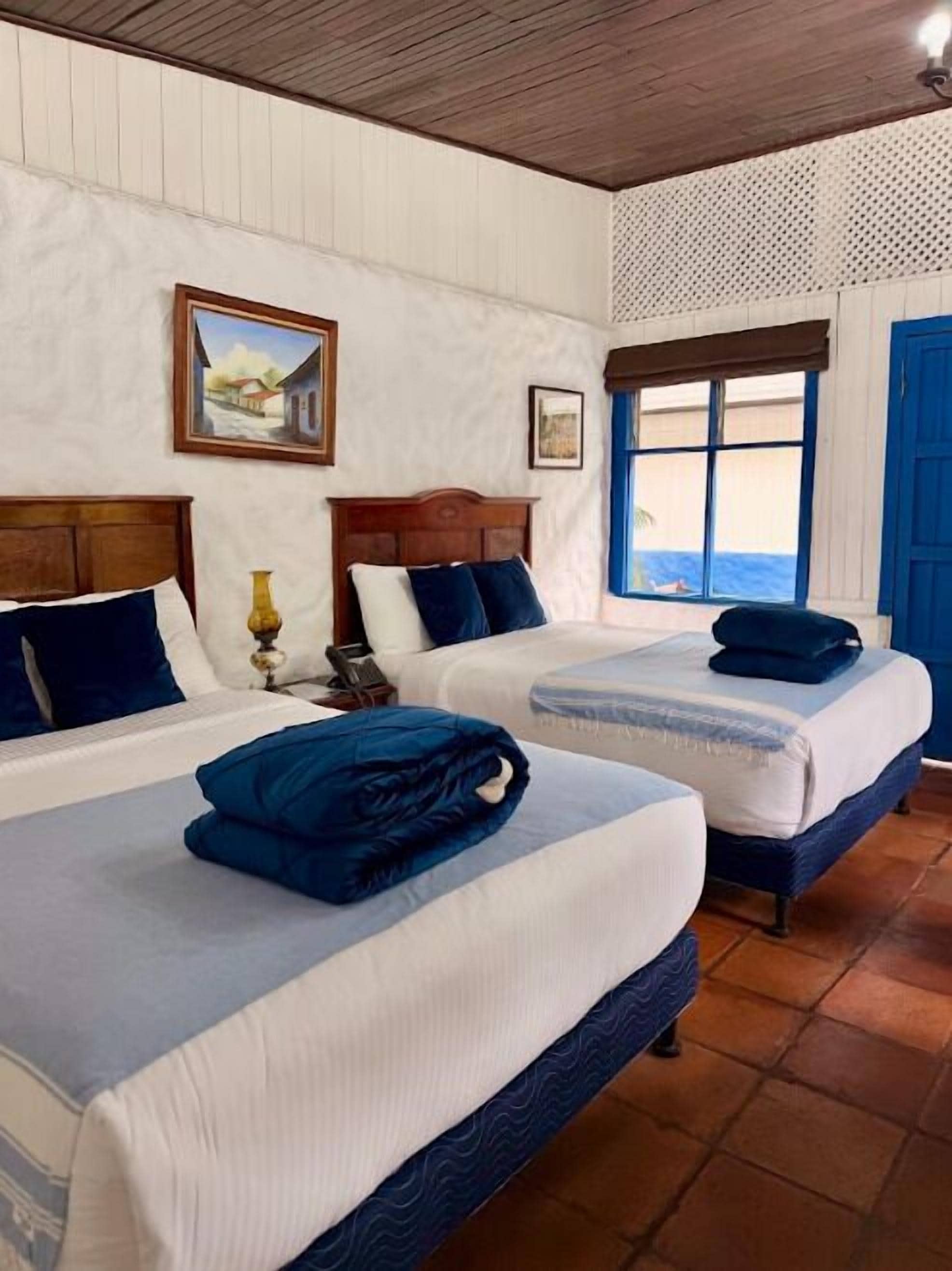 Habitación de hotel con dos camas, cabeceros de madera, una ventana con contraventanas azules y un cuadro en la pared.