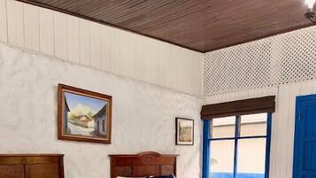 Habitación de hotel con dos camas, cabeceros de madera, una ventana con contraventanas azules y un cuadro en la pared.