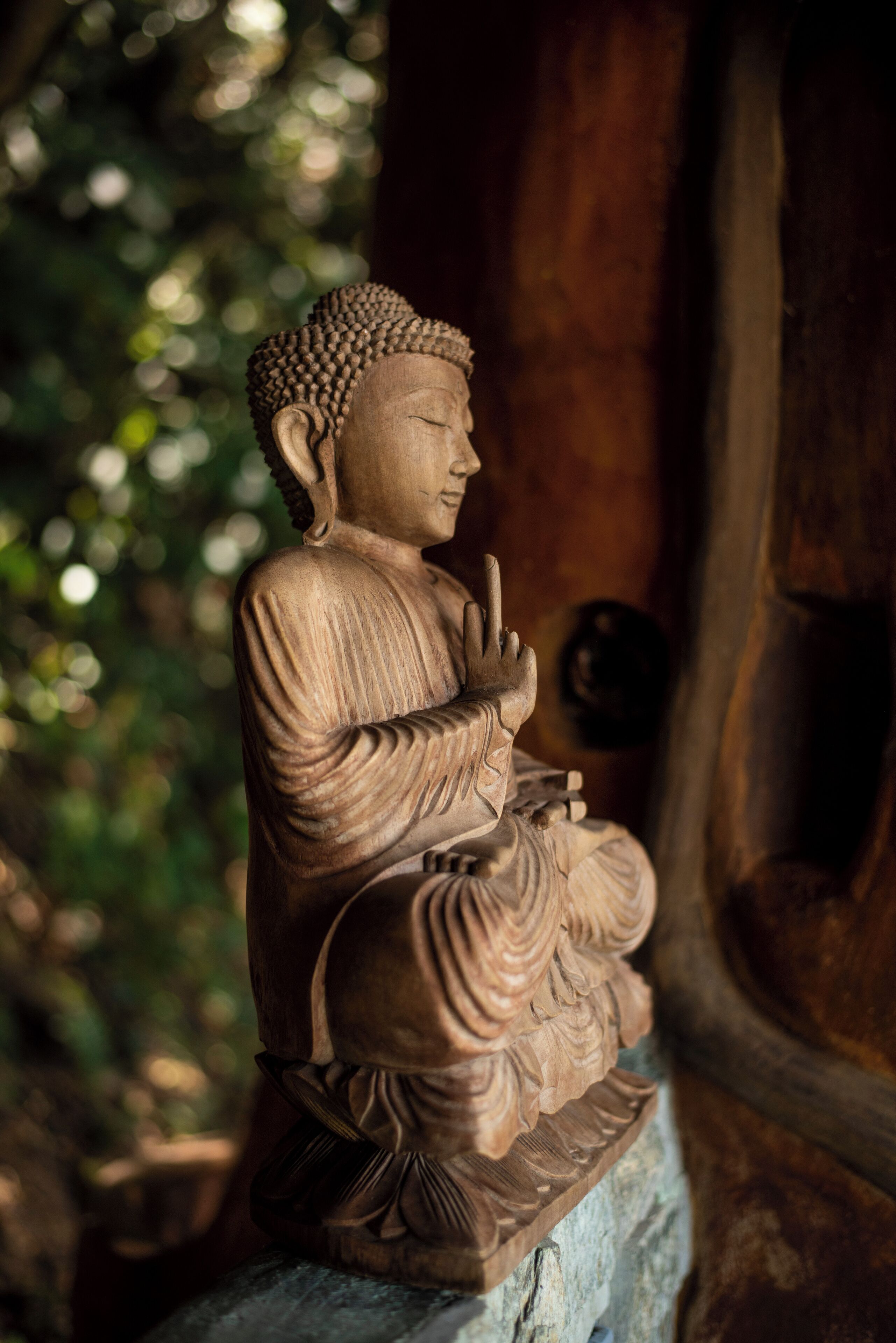 Una estatua sentada de una figura en pose meditativa, probablemente representando una figura religiosa o cultural, con un fondo de vegetación desenfocada.