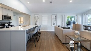 A modern kitchen with white cabinets, a central island, and bar stools. There is a dining area with a table and chairs, and a living room with a sofa and a TV. - **Family Retreat w/ Lake FRONT View (4bdr) (DeKalb County)