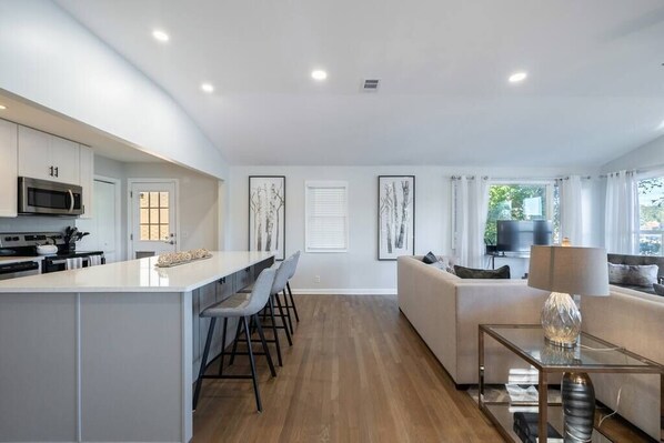 A modern kitchen with white cabinets, a central island, and bar stools. There is a dining area with a table and chairs, and a living room with a sofa and a TV. - Family Retreat w/ Lake FRONT View 4bdr (DeKalb County)
