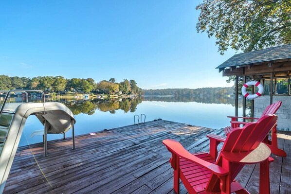 Un muelle de madera con sillas Adirondack rojas y un flotador salvavidas, con vista a un lago tranquilo y un cielo despejado.