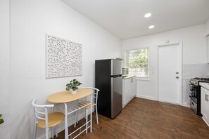 Een kleine keuken met een koelkast, een eettafel met stoelen en een raam met jaloezieën.