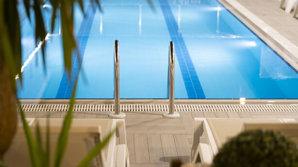 Una piscina con agua azul cristalina, rodeada por una terraza de madera y sillas de descanso blancas.