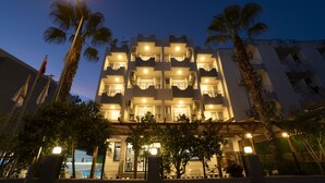 Un hotel de varios pisos con balcones, una entrada bien iluminada y palmeras.