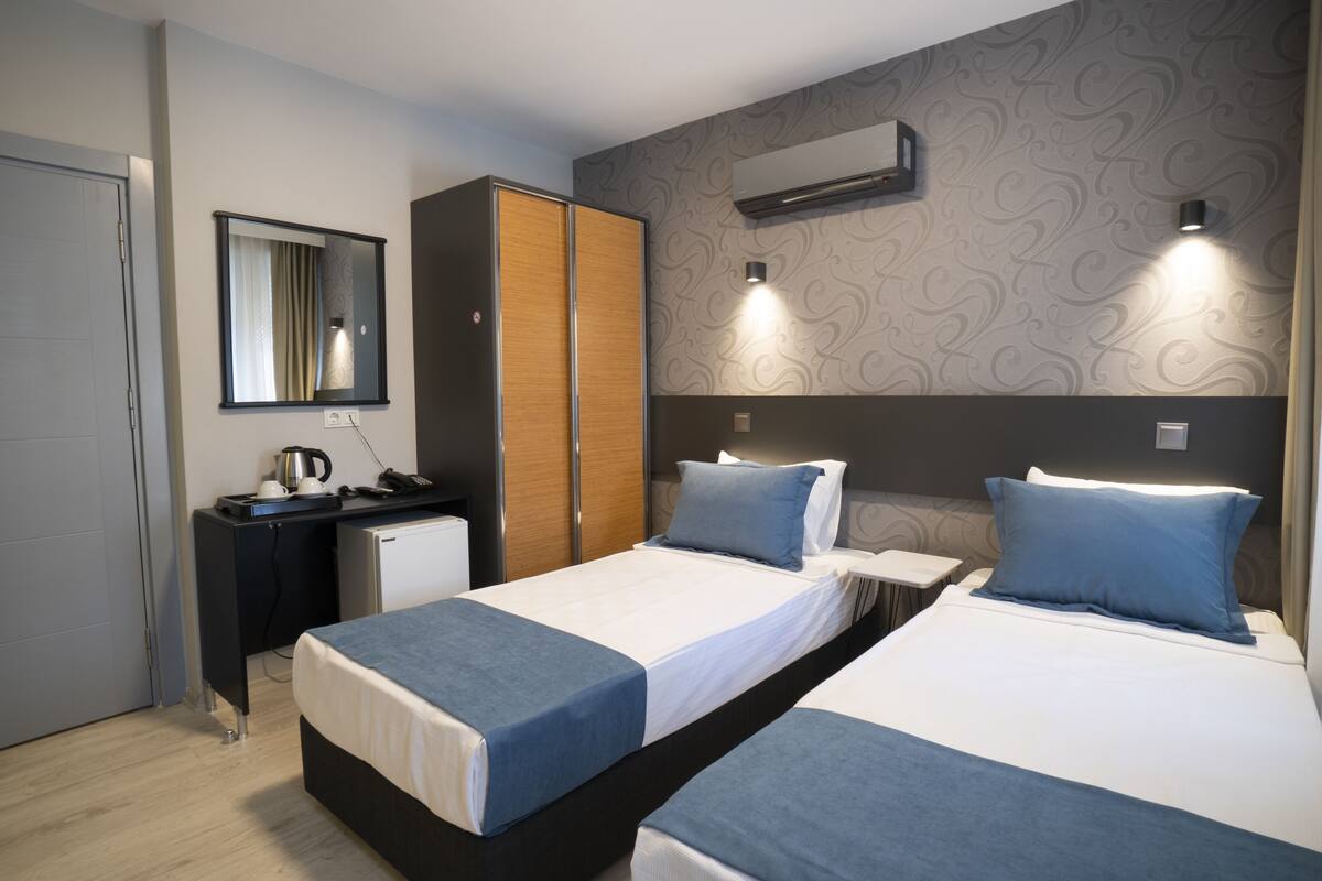 A hotel room with two beds, a desk, a wardrobe, a mirror, and an air conditioning unit.