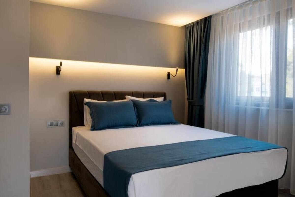 A bedroom with a bed, a headboard, a bedside table, a wall-mounted light, and a window with curtains.