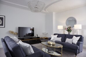 Un salon moderne avec un canapé bleu foncé, une table basse en verre, un téléviseur à écran plat, un lustre et un grand miroir rond accroché au mur.