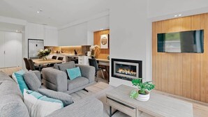 Ett modernt vardagsrum med en öppen spis, grå soffor, en matplats och ett kök i bakgrunden.