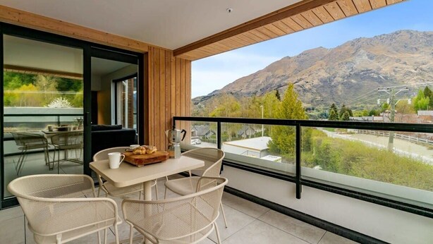 A balcony with a table and chairs, a view of mountains, and a modern interior with wooden beams and glass doors. - Coronet Bliss at La Residence du Parc (Queenstown)